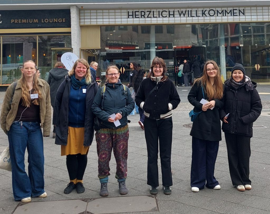 Gruppe von sechs Frauen steht vor einem Gebäude mit der Aufschrift 'HERZLICH WILLKOMMEN'. Alle tragen unterschiedliche Outfits und lächeln in die Kamera.