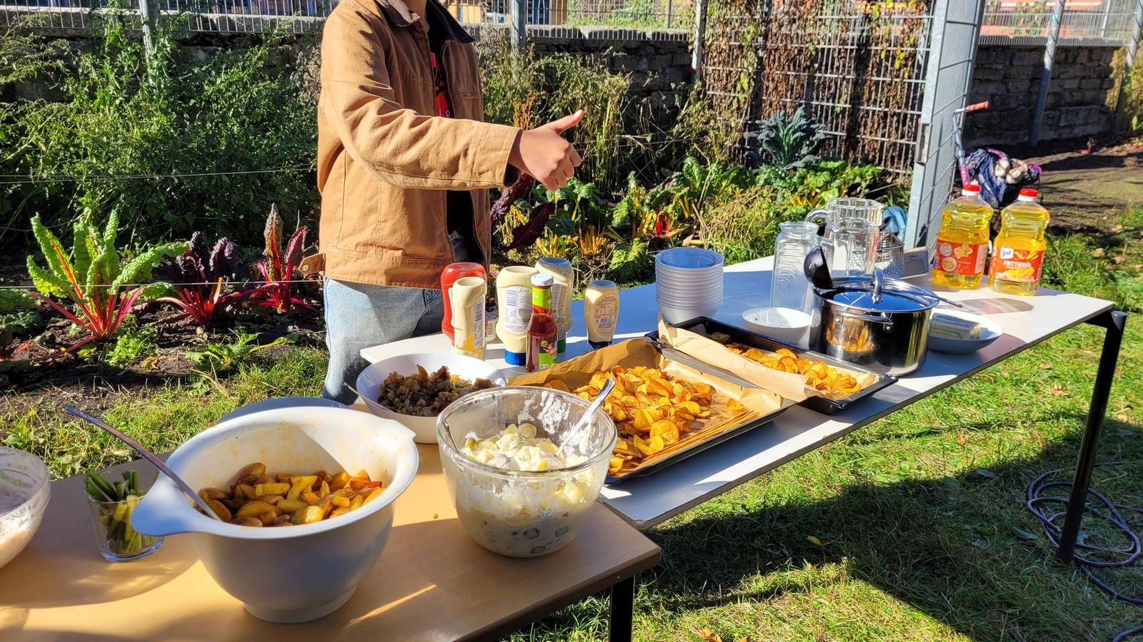 Tisch mit verschiedenen Schalen und Tellern mit Kartoffelgerichten und Saucen, im Hintergrund eine Person mit brauner Jacke, Garten mit Pflanzen