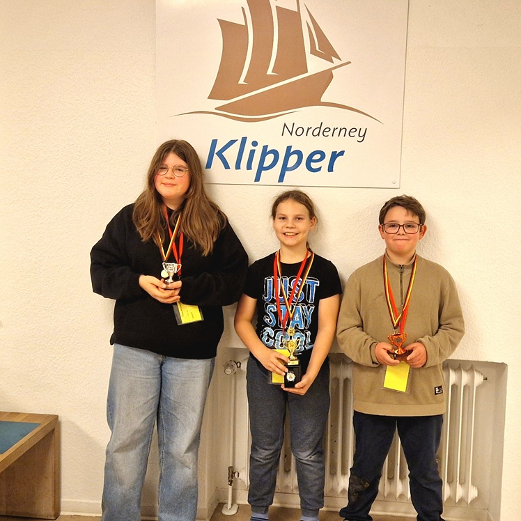 Drei Jugendliche mit Medaillen und Pokalen stehen vor einer Wand mit dem Logo 'Norderney Klipper'.