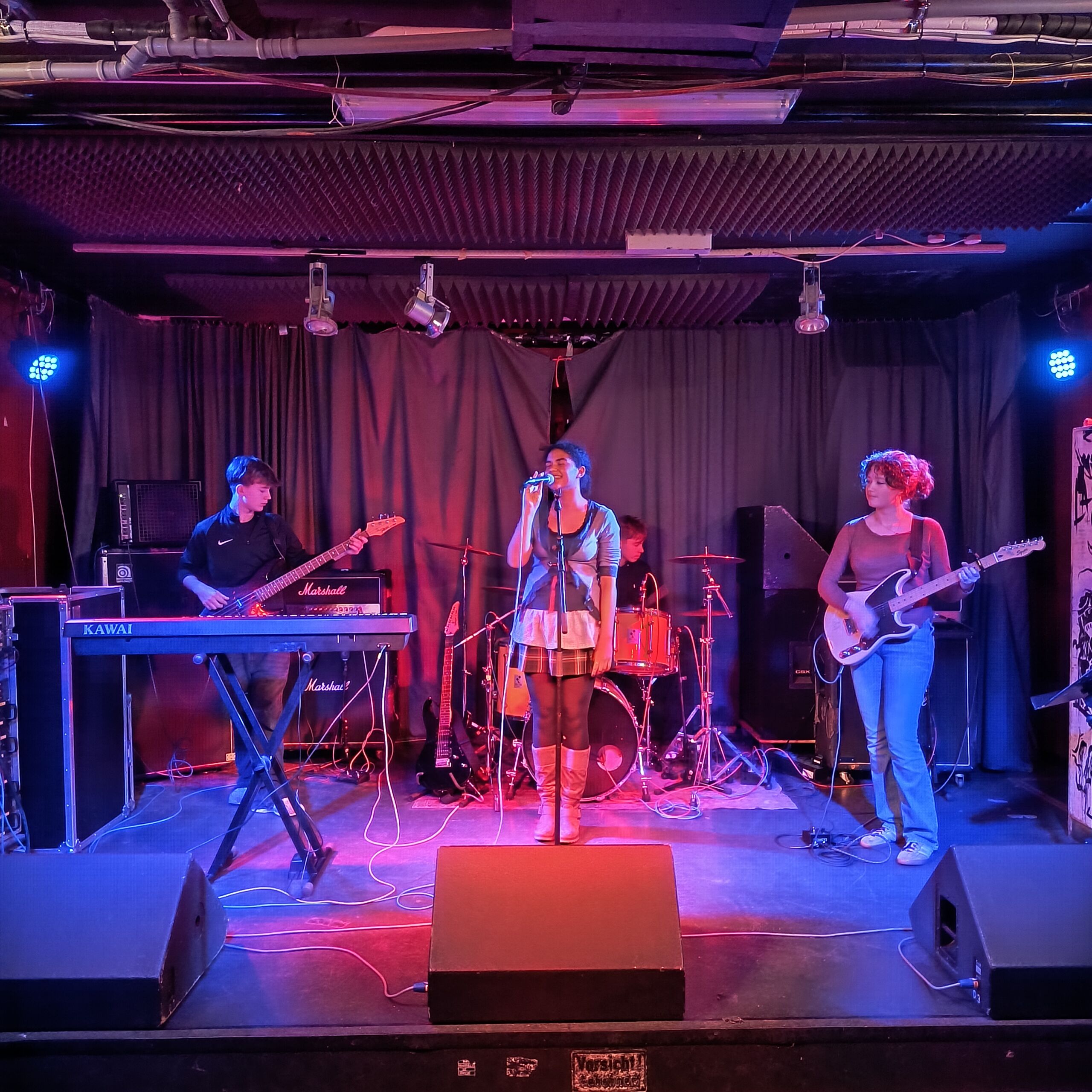 Vier Musiker auf einer Bühne: Sängerin mit Mikrofon in der Mitte, links Keyboardspieler, rechts Bassistin, hinten Schlagzeuger, bunte Bühnenbeleuchtung.