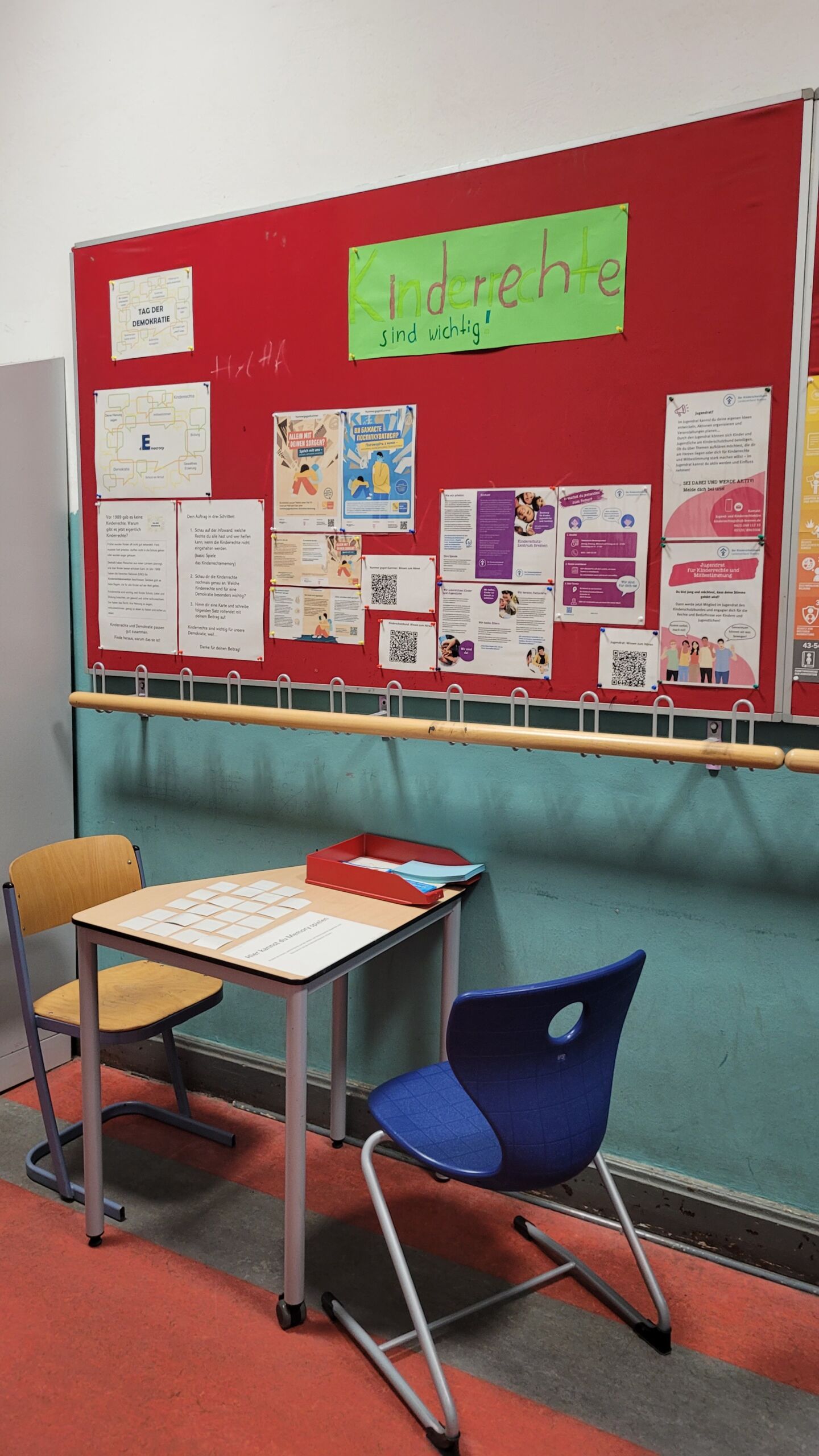 Die _Station E__ Kinderrechte sind wichtig! Roter Pinnwand mit verschiedenen Informationsblättern und einem grünen Plakat mit der Aufschrift 'Kinderrechte sind wichtig!' über einem kleinen Tisch mit zwei Stühlen in einem Klassenraum der Gesamtschule Bremen Mitte.