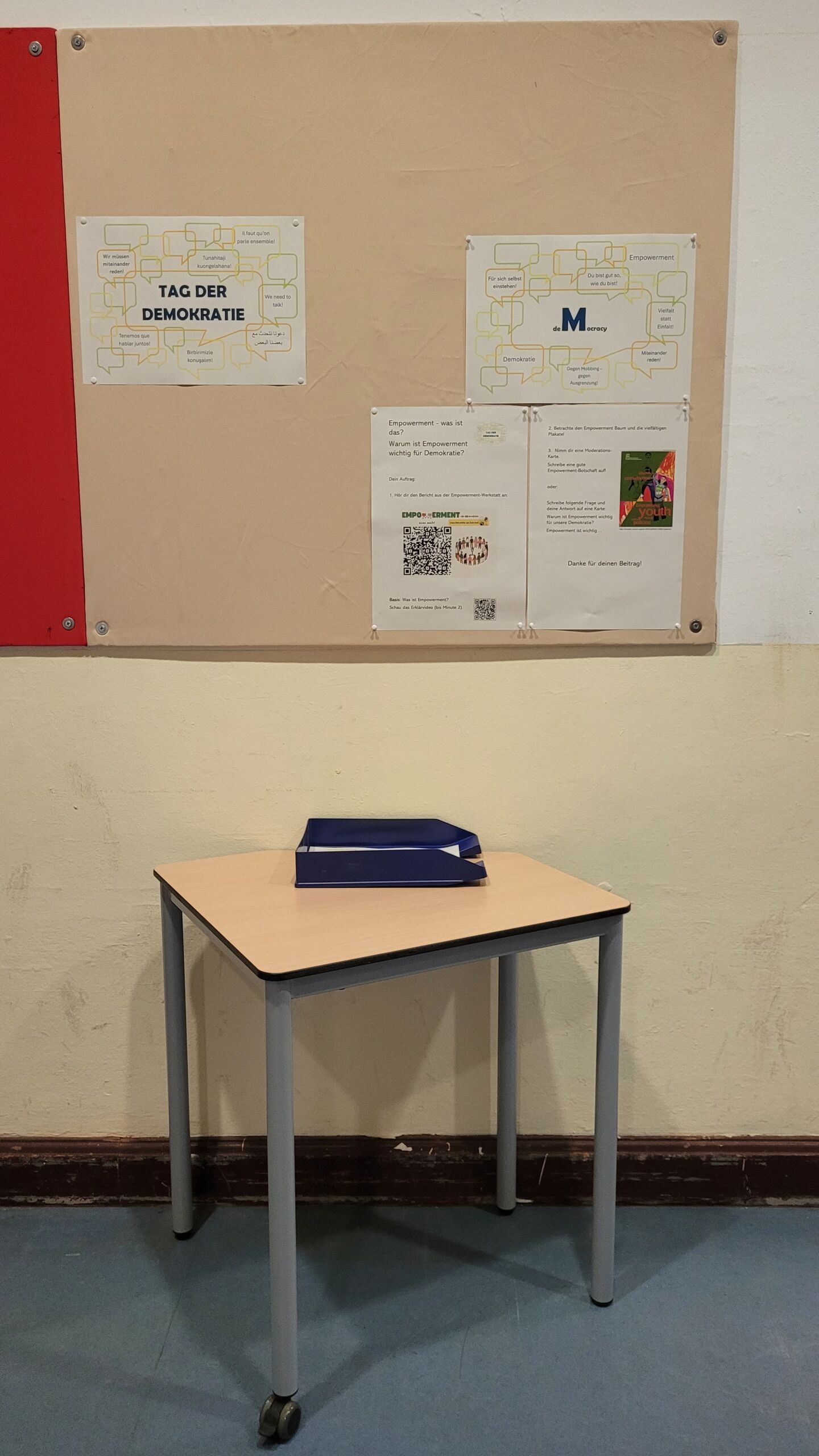 Die _Station M__ Empowement Schlichter Tisch mit zwei blauen Ordnern vor einer Pinnwand mit vier Aushängen zur Station M des Tages der Demokratie 2025 an der Gesamtschule Bremen Mitte.