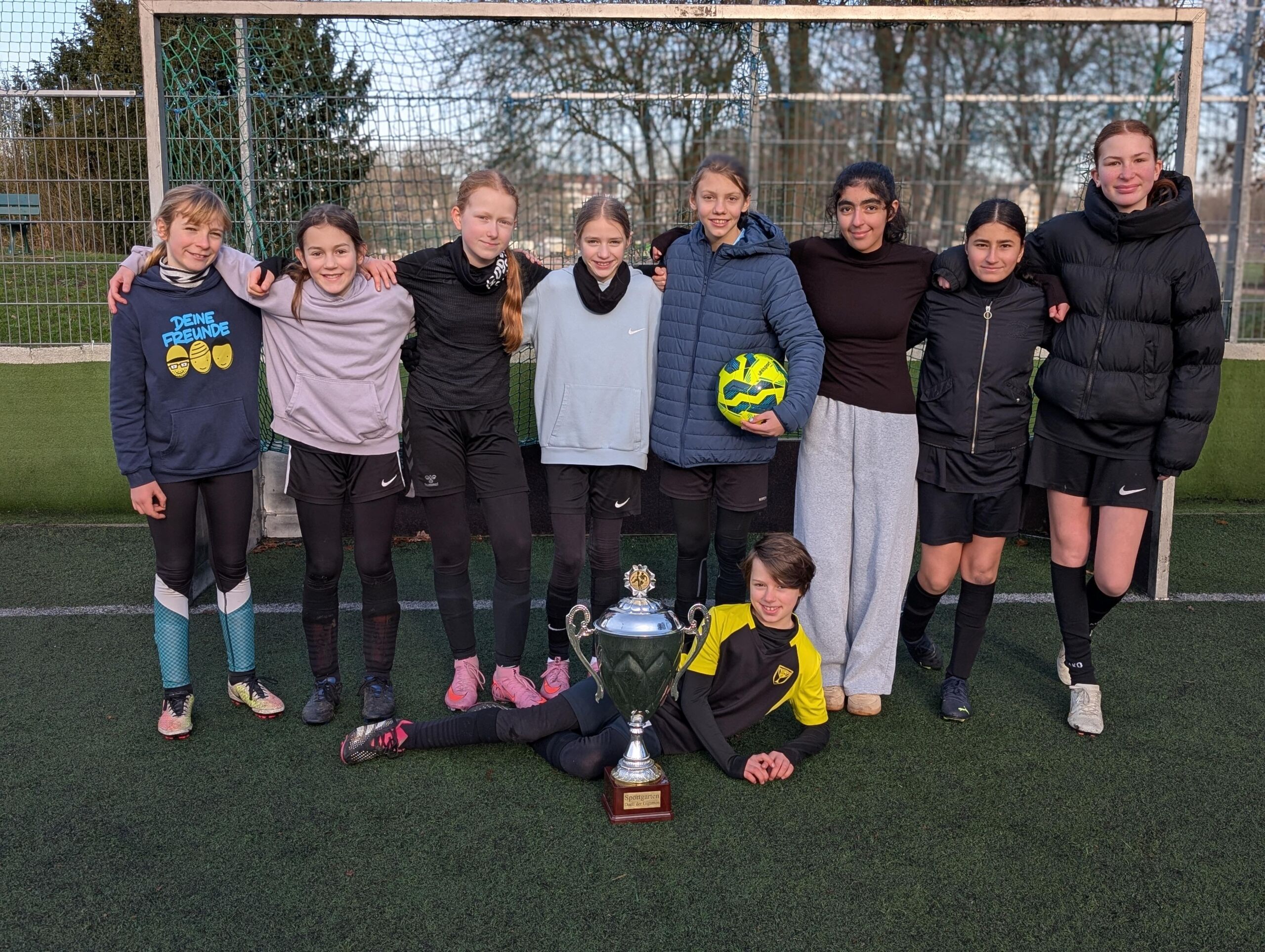 Mädchenteam der Gesamtschule Bremen Mitte steht in Sportkleidung auf einem Fußballfeld, ein Spieler liegt vor einer großen Trophäe, eine weitere hält einen Fußball