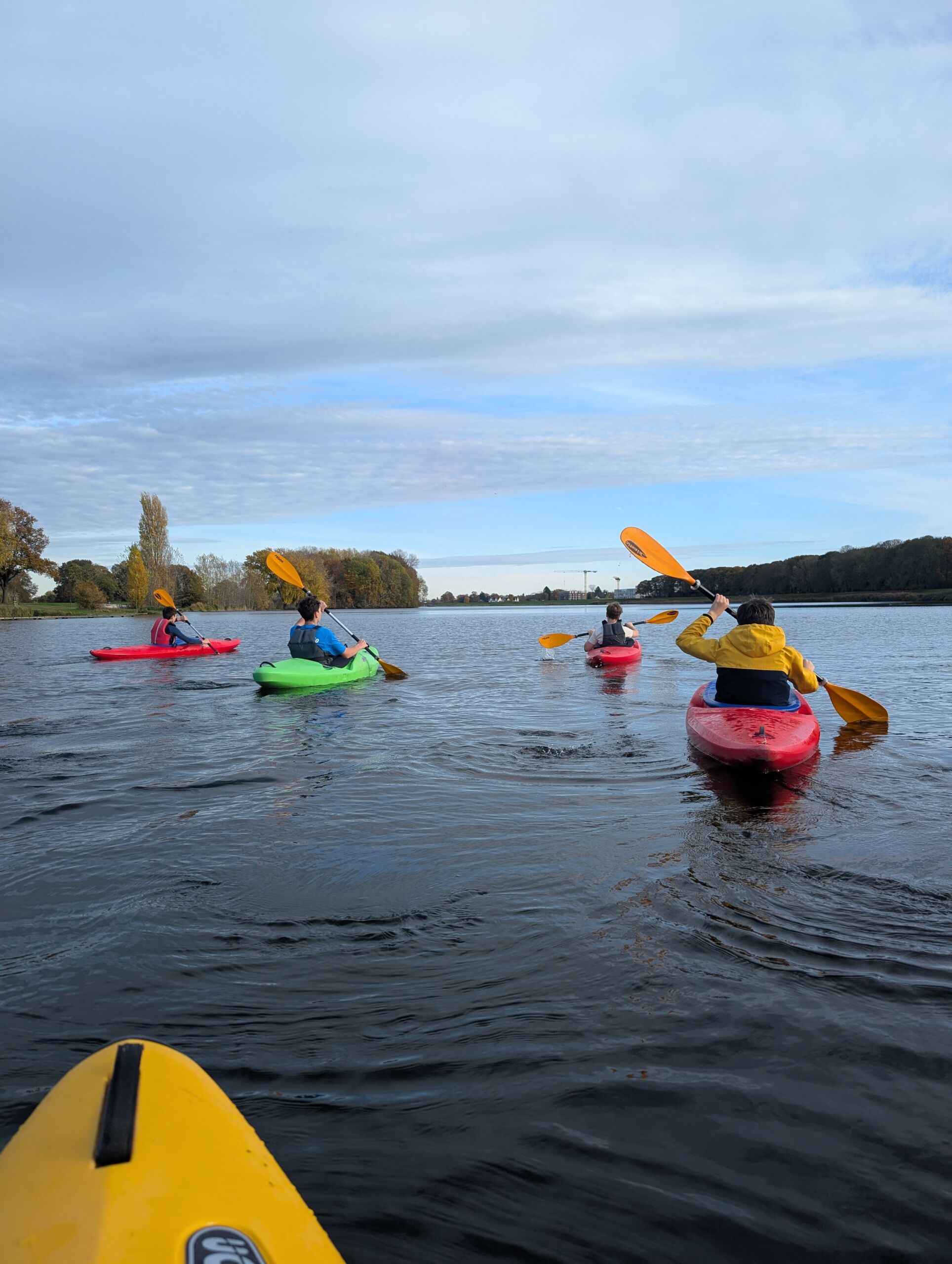 Vier Personen paddeln in bunten Kajaks auf einem ruhigen Gewässer, aufgenommen aus der Perspektive eines fünften Kajaks