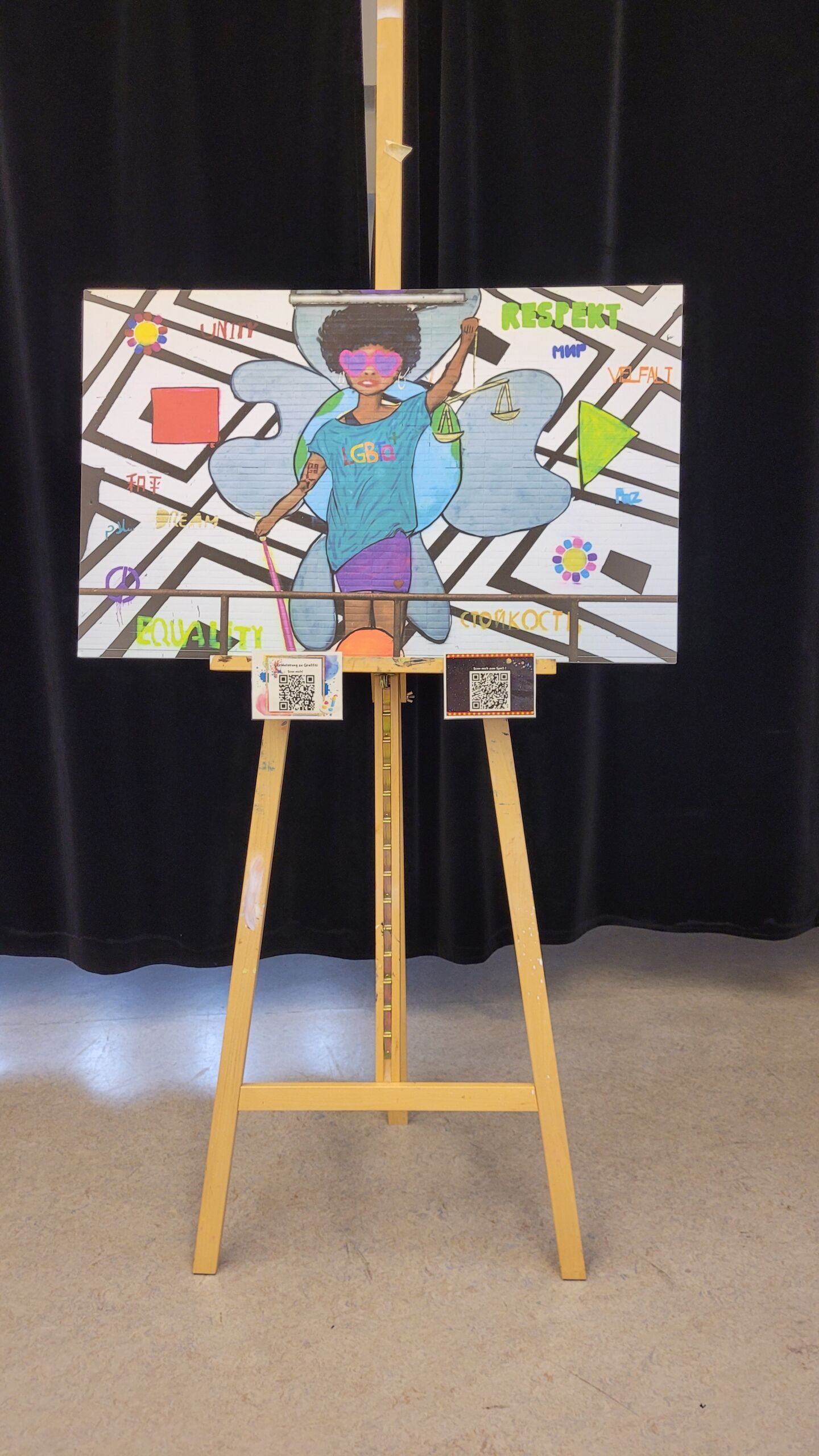 _Station D__ Kunst von der Welterbe-Tagung 2025 – Das Graffiti _Justicia_ Gemaltes Graffiti mit einer stehenden Person vor abstraktem Hintergrund, umgeben von bunten geometrischen Formen und den Worten 'EQUALITY', 'RESPECT' und 'JUSTICIA', präsentiert auf Staffelei vor schwarzem Vorhang.