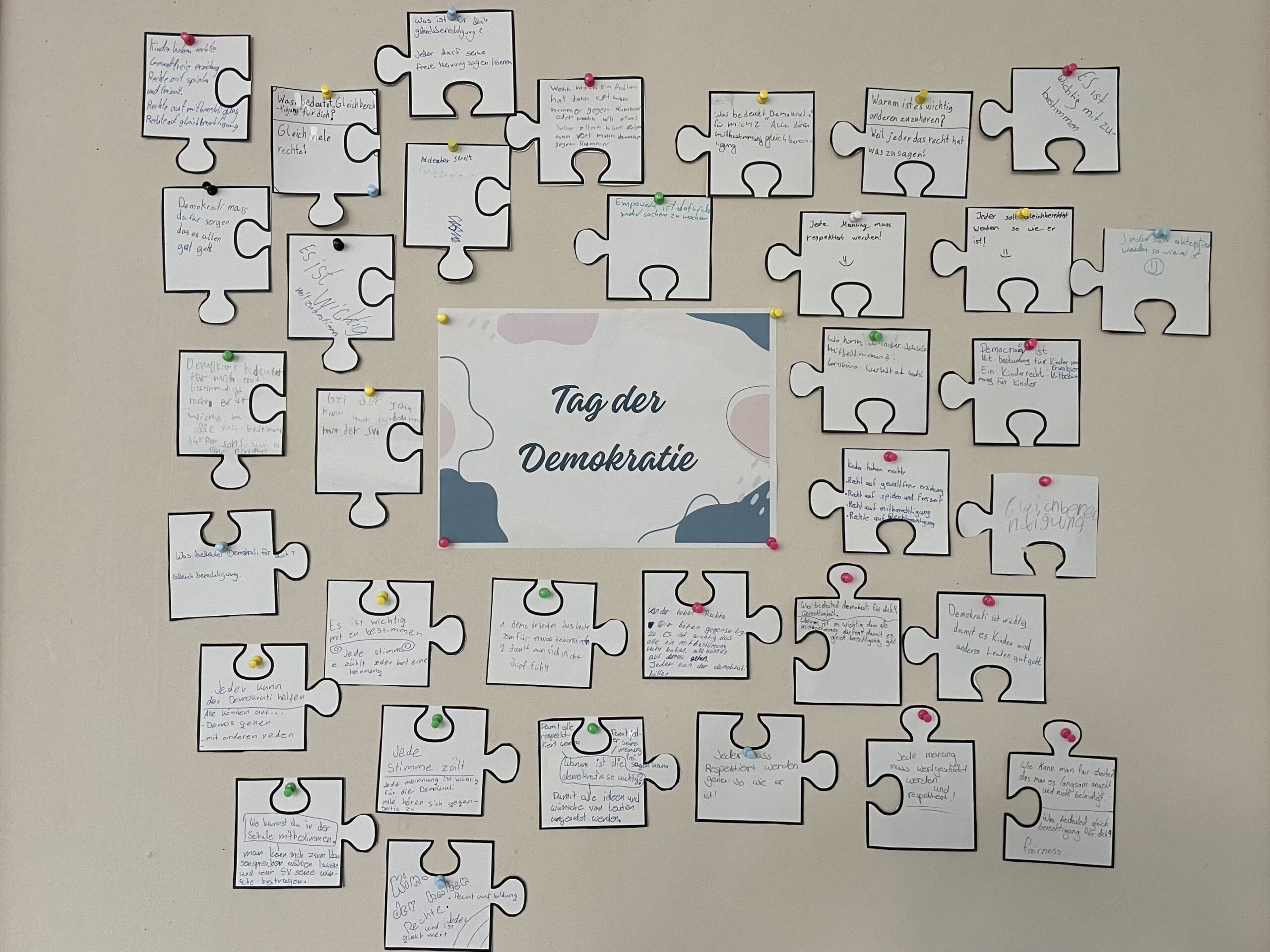 Stationen-Beiträge von Schüler_innen aus der Werkstatt _Von der Diktatur zur Demokratie_ Wand mit Puzzleteilen aus Papier, die Schülerbeiträge zum Tag der Demokratie 2025 an der Gesamtschule Bremen Mitte zeigen, mittig ein Schild mit der Aufschrift 'Tag der Demokratie'.