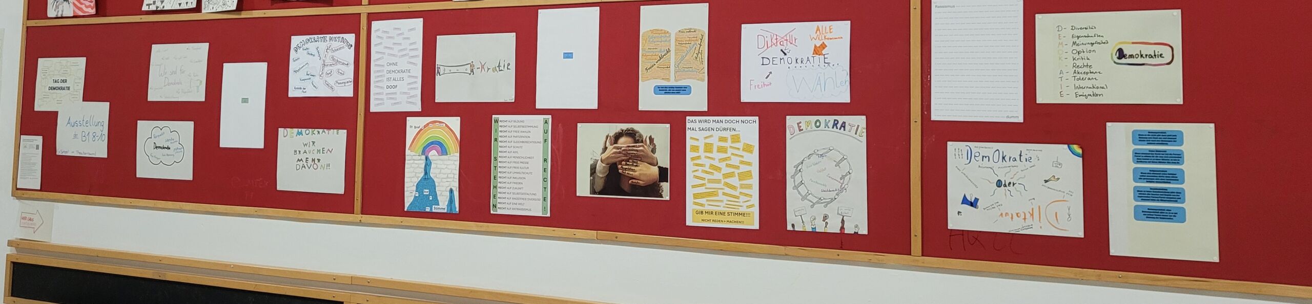 Stationen-Beitrag der B1 (8-10) Plakate zu _Demokratie_ Wandtafel mit mehreren handgeschriebenen und gezeichneten Plakaten zum Thema Demokratie, erstellt von Schülerinnen und Schülern der Klasse B1 (8-10) der Gesamtschule Bremen Mitte für den Tag der Demokratie 2025.