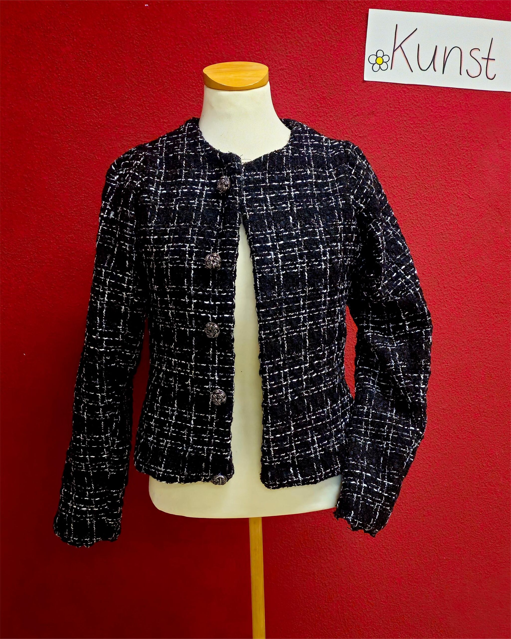 Schwarze Chanel-Jacke mit weißem Karomuster auf Schaufensterpuppe vor roter Wand mit handgeschriebenem Schild 'Kunst'