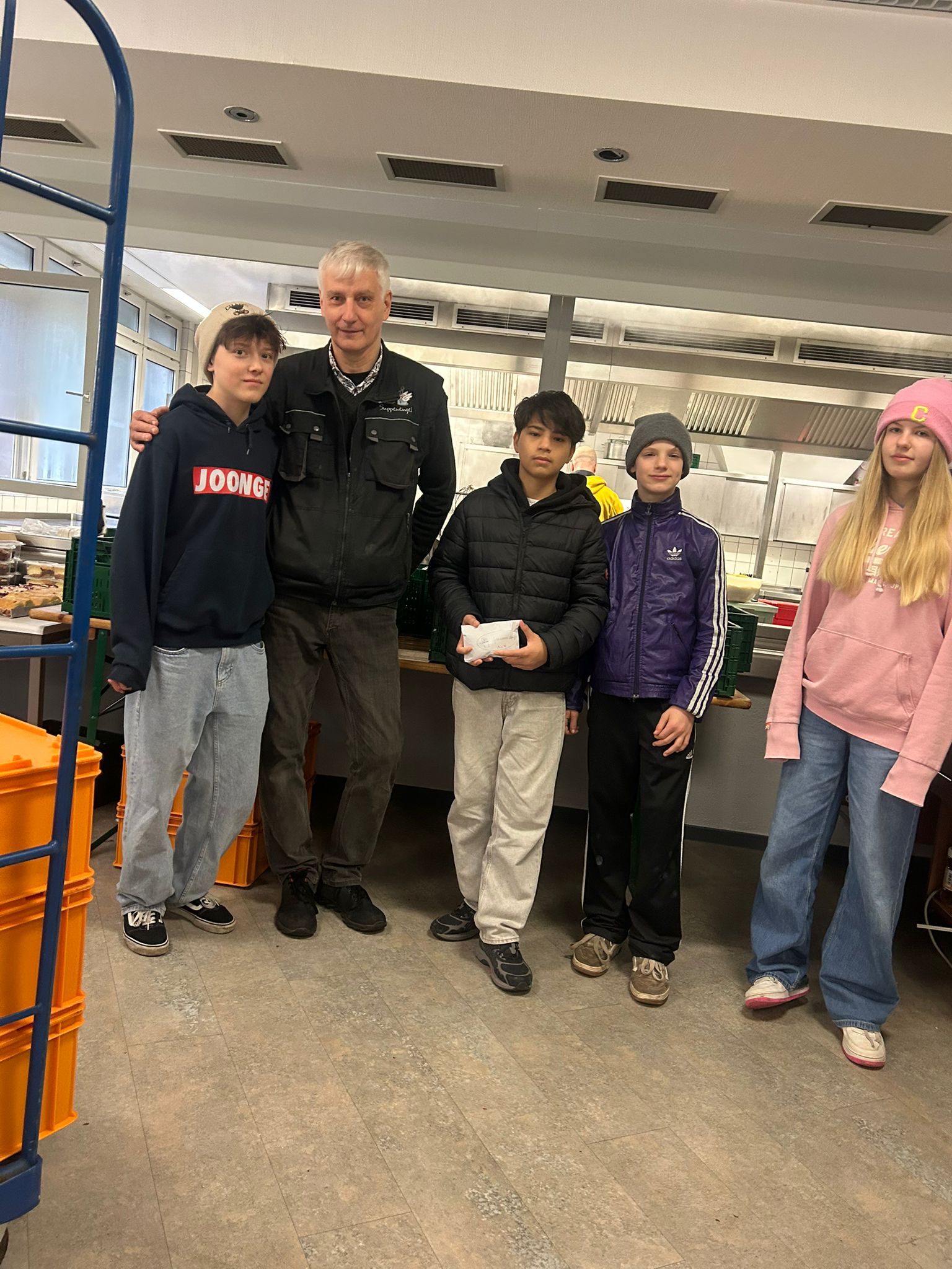 IMG-20260310-WA0000 Fünf Personen stehen in einer Küche, darunter vier Jugendliche in Freizeitkleidung und eine erwachsene Person in schwarzer Jacke, eine Person hält einen Umschlag