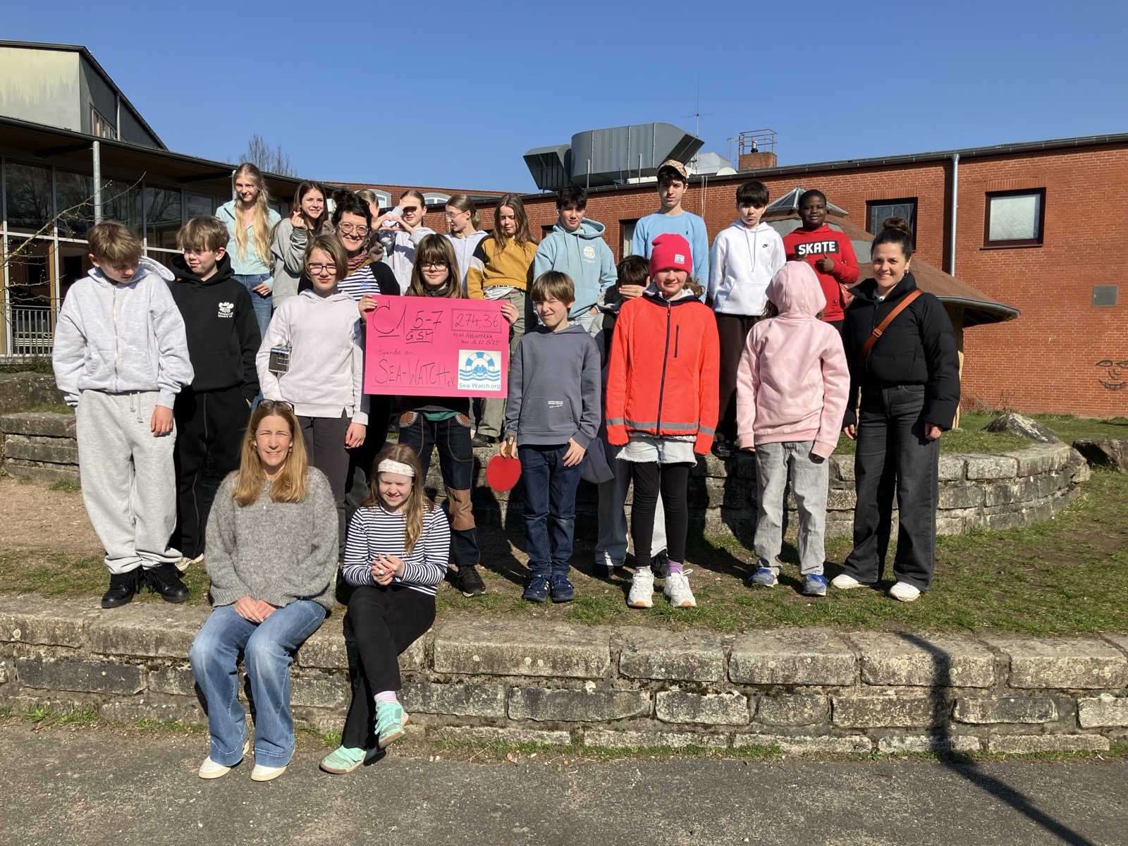 Gruppe von Jugendlichen und Erwachsenen steht auf einer steinernen Treppenanlage vor einem roten Backsteingebäude, eine Person hält ein pinkes Plakat mit handgeschriebenem Text und einem gedruckten Logo