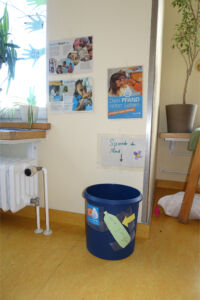 Blauer Behälter mit Aufklebern und einem Papierzettel mit der Aufschrift 'Spende die Hand' an einer Wand mit mehreren Informationsplakaten in einem Raum mit gelbem Boden und Pflanzen auf der Fensterbank und einem Holzregal.