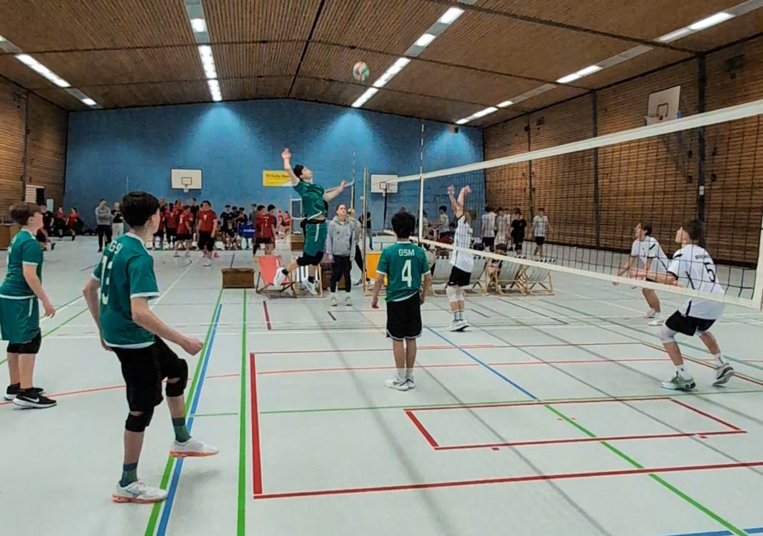Jugendliche Volleyballspieler in Sportkleidung beim Spiel in einer Halle mit Holzwänden und Basketballkörben im Hintergrund