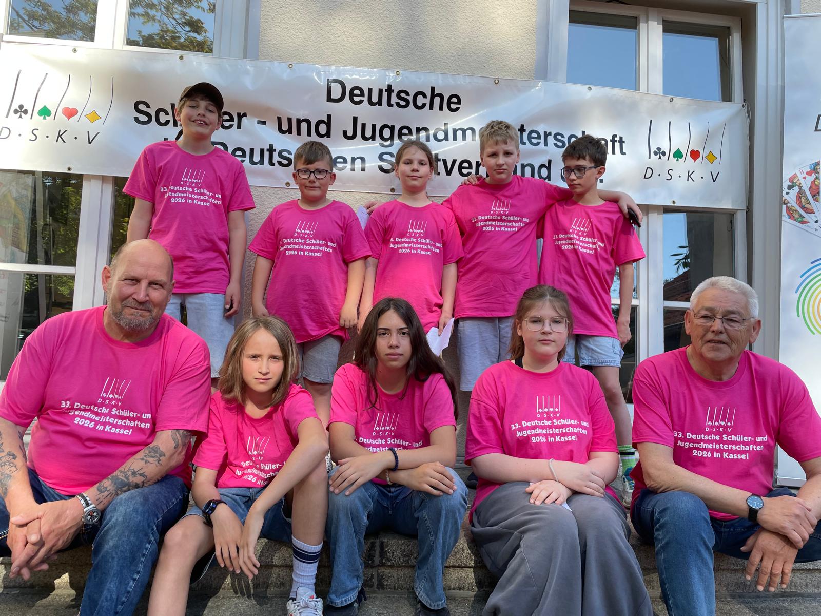 Gruppe von zehn Personen in pinken T-Shirts mit Aufdruck zur 33. Deutschen Schüler- und Jugend-Meisterschaft 2026 in Kassel vor einem Banner mit gleichem Veranstaltungstitel