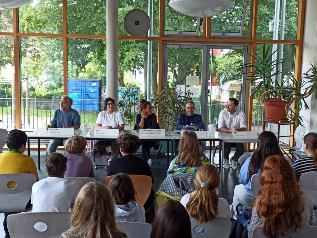 Fünf Personen sitzen an einem langen Tisch vor großen Fenstern, vor ihnen sitzen mehrere Schüler auf Stühlen, im Raum stehen große Pflanzen.