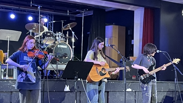 Schülerband mit Violine, Akustikgitarre, E-Bass und Schlagzeug auf einer Bühne bei einem Auftritt