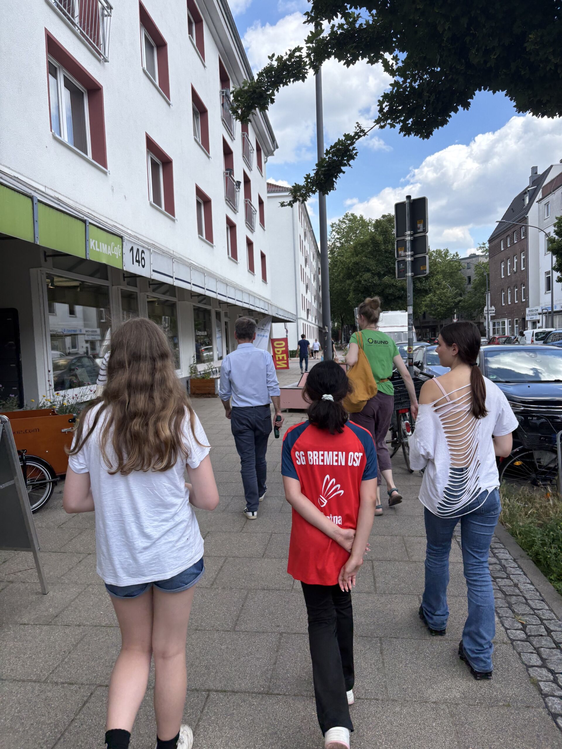 2026-06-18_ExkursionKlimazoneFindorff2_c_Martje_Behrens Gruppe von Menschen, darunter ein Kind mit rotem T-Shirt mit Aufdruck 'SG BREMEN OST', geht auf einem Bürgersteig an einem mehrstöckigen Gebäude mit der Nummer 146 entlang.