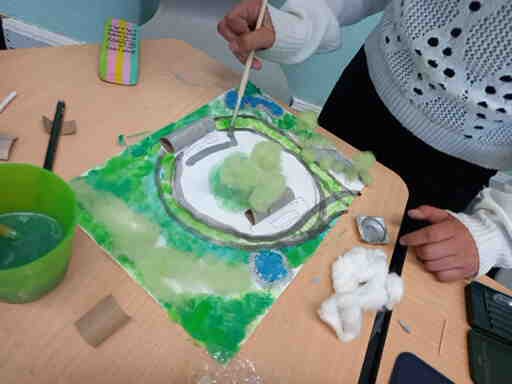 2026-KlimaanpassungHaltestelleWeserwehr_Aatna&Nele1_c_H_Oelgeklaus Hand einer Person in weißem Pullover mit schwarzen Punkten, die mit einem Pinsel an einem Modell mit grüner und blauer Farbe auf einem Tisch arbeitet