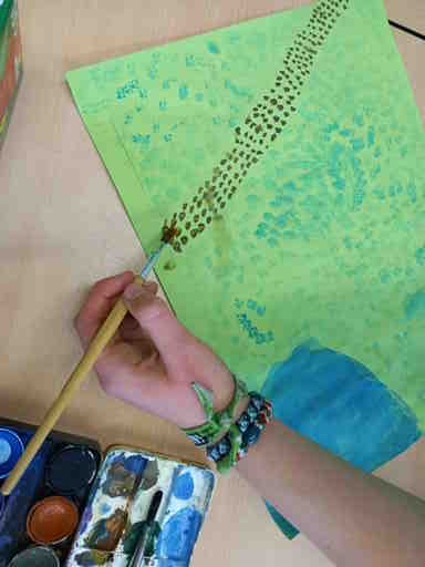2026-KlimaanpassungTermitenhaus_Jonna2_c_H_Oelgeklaus Hand malt mit Pinsel und brauner Farbe eine Reihe von Punkten auf grünes Papier, um eine Termitenstraße darzustellen