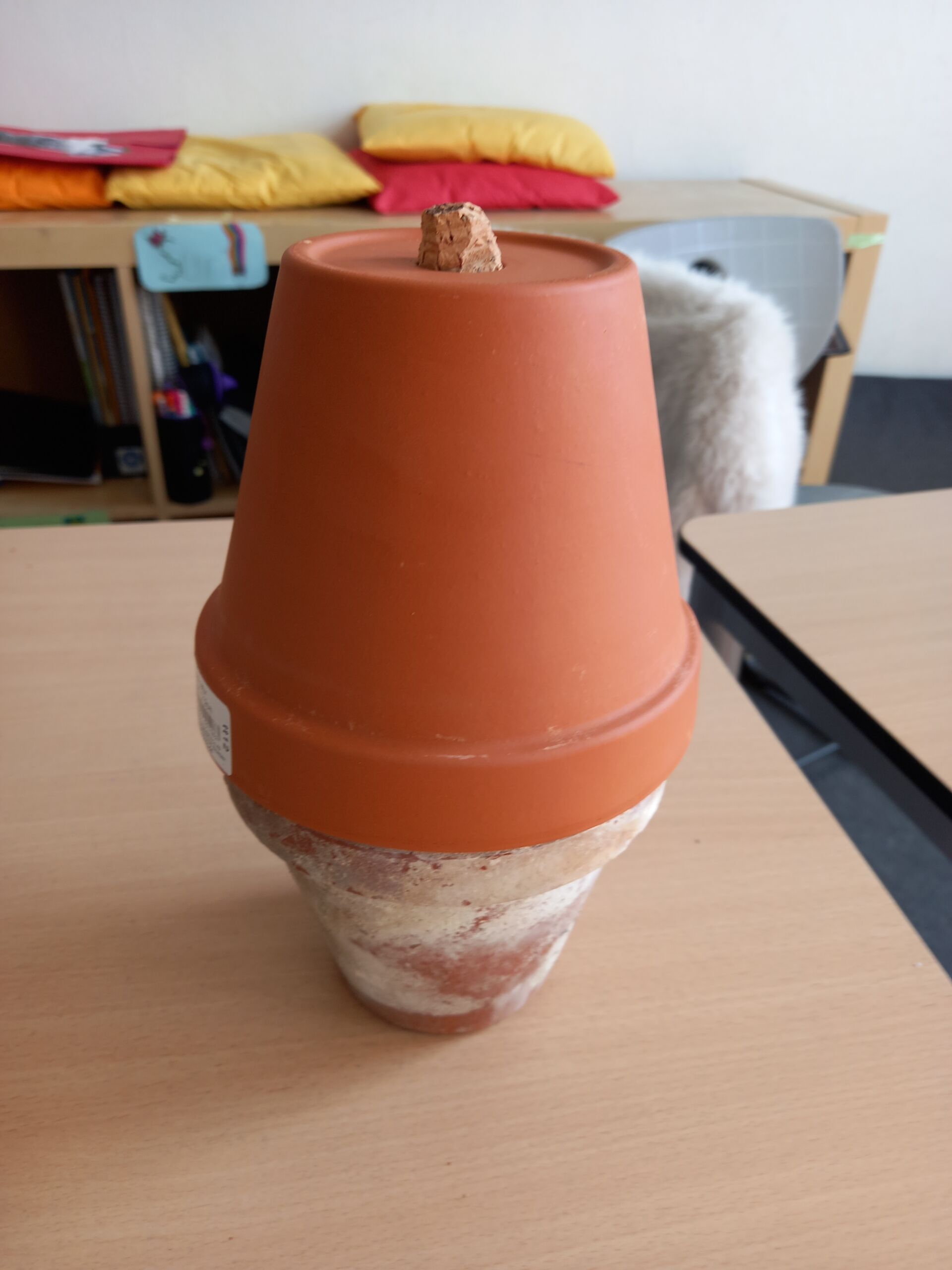 2026_OllaImKlassenzimmer_c_H_Oelgeklaus Zwei ineinander gestapelte Tontöpfe mit einem Korken auf dem oberen Topf stehen auf einem Tisch in einem Klassenraum.